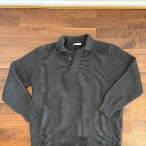 Mörkgrå stickad pikétröja från Zara - Ribbstickad mörkgrå pikétröja från Zara med klassisk krage och knappar framtill. Tröjan har lång ärm och är tillverkad i ett mjukt stickat material som ger en chill och stilren vibe.