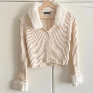 Beige ribbad kofta med fuskpälsdetaljer - Superfin beige ribbad kofta från SHEIN med fluffig fuskpäls på krage och ärmslut. Koftan har knappar framtill, croppad passform och långa ärmar. Perfekt för dig som vill ha en mysig och trendig look.