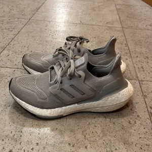 Grå Adidas Ultraboost sneakers - Säljer ett par grå Adidas Ultraboost sneakers med stickad ovandel och klassiska tre ränder på sidan. Skorna har en vit, dämpad sula och snörning framtill. Perfekta för dig som gillar stilrena och bekväma sneakers med sportig känsla.