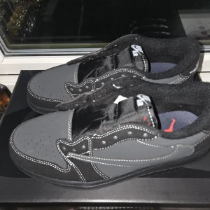 Nike Air Jordan 1 Low Travis Scott - Säljer ett par Nike Air Jordan 1 Low Travis Scott i svart och grått med vita kontrastsömmar. Skorna har en omvänd swoosh, broderad bi på hälen och klassisk Jordan-logga på innersulan. Materialmix av mocka och syntet, med platt sula och rund tå.