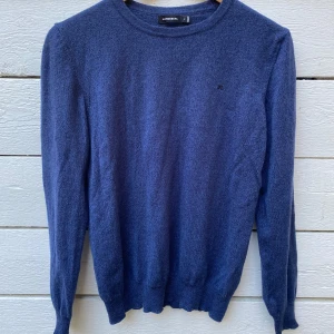 Blå stickad tröja från J.Lindeberg - blå stickad tröja från J.Lindeberg i storlek M. Tröjan har rund hals och långa ärmar, med diskret JL-logga på bröstet. Perfekt för dig som gillar stilrena och klassiska plagg med skön passform.