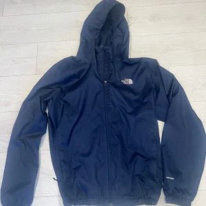 Marinblå vindjacka från The North Face - Snygg marinblå vind/regn jacka från The North Face med huva och hel dragkedja framtill. Jackan har meshfoder, elastiska muddar och justerbar nederkant med snörning. Perfekt för blåsiga dagar och outdoor-aktiviteter. Logga på bröstet och på baksidan, sparsamt andvänd inga defekter