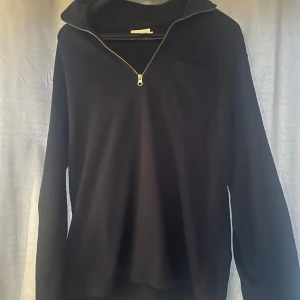 Svart half zip-tröja från East West - Svart långärmad tröja från East West med half zip-dragkedja och hög krage. Tröjan har en bröstficka och är tillverkad i mjukt bomullsmaterial. Perfekt för dig som gillar en clean och stilren look.