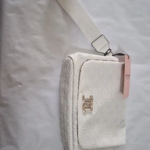 Vit axelväska från Juicy Couture - Snygg vit axelväska från Juicy Couture med präglat JC-mönster över hela väskan och glittrande JC-logga framtill. Väskan har bred axelrem och innerfoder med Juicy Couture-tryck. Perfekt för dig som vill ha en trendig och stilren accessoar.