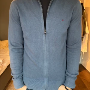 Blå kofta från Tommy Hilfiger M - Snygg blå kofta från Tommy Hilfiger i pima bomullsblandning med kashmir. Tröjan har halv dragkedja, ribbade muddar och klassisk Hilfiger-logga på bröstet. Ståkrage med röd, vit och marinblå rand på insidan ger en extra detalj.