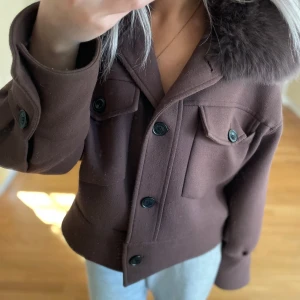 Brun jacka med pälskrage från Meotine - Säljer en brun jacka från Meotine med avtagbar pälskrage. Jackan har stora bröstfickor med lock, knappar framtill och en boxig, lite croppad passform. Materialet är ullmix och kragen är i mjuk päls. Perfekt för höst och vinter!