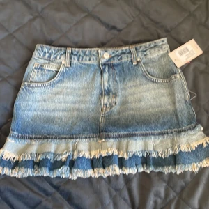 Blå jeanskjol med volang från Nelly - Säljer en blå jeanskjol från Nelly med volangkant och fransade detaljer längst ner. Kjolen har klassisk femficksdesign, bälteshällor och knäppning fram. Perfekt för dig som gillar denim med en twist och vill sticka ut lite extra. Aldrig använd som du kan se på prislappen. Köpt för 600 säljer för 450. 