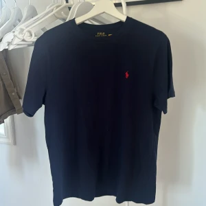 Mörkblå t-shirt från Polo Ralph Lauren - Snygg mörkblå t-shirt från Polo Ralph Lauren i storlek XL (känns som i storlek M). Klassisk design med rund hals och korta ärmar, tillverkad i mjuk bomull. På bröstet finns det ikoniska röda Polo-logot broderad. Perfekt för dig som gillar stilrena och tidlösa plagg. 