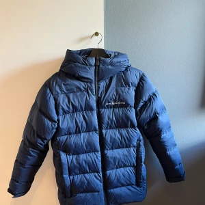 Blå pufferjacka från Sail Racing - Säljer en blå pufferjacka från Sail Racing med huva och dragkedja. Jackan har en quiltad design och logga på bröstet samt på huvan. Perfekt för kalla dagar och riktigt snygg till streetwear-stilen.