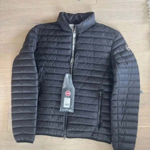 Svart dunjacka från Colmar - Snygg svart dunjacka från Colmar med horisontella sömmar och hög krage. Jackan har dragkedja framtill, två sidofickor med dragkedja och Colmar-logga på ärmen. Perfekt för kyliga dagar och riktigt clean look.