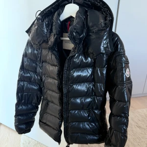 Svart pufferjacka från Moncler - Säljer en svart pufferjacka från Moncler med glansig finish och klassisk logga på ärmen. Jackan har huva, dragkedja framtill och sidofickor med dragkedja. Perfekt för kalla dagar och har en riktigt snygg streetstil.