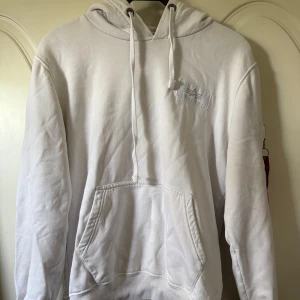 Vit hoodie från Alpha Industries - Vit hoodie från Alpha Industries med stor svart logga på ryggen och mindre logga på bröstet. Tröjan har huva med snörning, magficka och en cool ficka med dragkedja på ärmen. Mjuk bomullskvalitet, har även nån fläck på fickan som knappt syns.