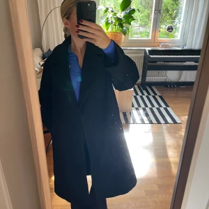 Svart kappa med klassisk krage - Säljer en svart kappa med klassisk krage och raka fickor. Jackan har en rak och loose passform. Perfekt för dig som vill ha en stilren och enkel look till höst och vinter. Materialet är ullblandning som håller dig varm. Jag har använt den som vinterjacka tidigare året. 