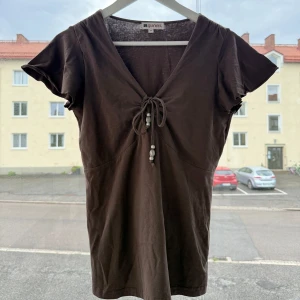 Brun topp med knyt och pärlor Gianin - Snygg brun topp från gianin med v-ringning och knyt framtill dekorerat med pärlor. Modellen har korta ärmar och är tillverkad i mjuk bomull. Känns som st xs/s figur sydd passform 
