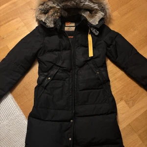 Svart dunjacka Parajumpers med päls - Svart lång dunjacka från Parajumpers med ikonisk gul rem och pälsfodrad huva. Jackan har flera fickor, snygga detaljer som metallspänne och patch på ärmen. Slim fit-modell, perfekt för kalla vinterdagar. Riktigt stilren och varm jacka med dragkedja och knappar.