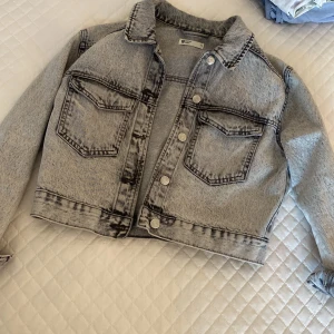 Grå croppad jeansjacka från Perfect Jeans - Säljer en grå croppad jeansjacka från Gina  i storlek XS. Jackan har tydliga svarta sömmar, två stora bröstfickor med lock och knappar framtill. Klassisk krage och långärmad modell, perfekt för att lyfta vilken outfit som helst.