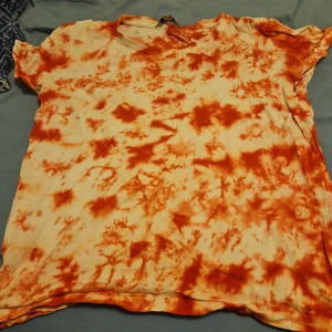 Orange tie-dye t-shirt från TIKI - Säljer en riktigt chill t-shirt från TIKI i storlek 170. Den har ett orange och vitt tie-dye mönster som verkligen sticker ut. T-shirten är kortärmad och gjord i mjuk bomull, perfekt för dig som gillar en avslappnad och färgstark stil.