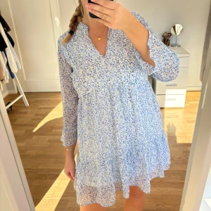 Blommig klänning  - Supersöt kort klänning från Pigalle i ljusblått och vitt med blommigt mönster. Klänningen har v-ringning och volangkant nertill. I storlek 36, använd fåtal gånger. 