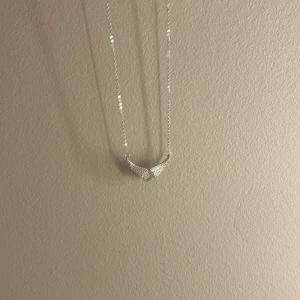 Halsband med vingar i silverfärg - Säljer ett snyggt halsband i silverfärg med ett hänge format som två vingar som möts i mitten. Kedjan är tunn och smidig, och vingarna har små glittrande stenar som ger extra bling. Perfekt accessoar för dig som gillar unika detaljer och stilren design. Dock lite defekt på baksidan som syns på bilden men märks inte på eftersom det är på bak sidan 