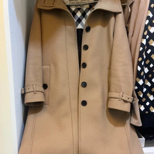 Beige trenchcoat från Burberry - Klassisk beige trenchcoat från Burberry med bälte i midjan, knappar framtill och snygga spänndetaljer vid ärmsluten. Fodret har det ikoniska rutmönstret. Perfekt för dig som vill ha en tidlös och stilren jacka. Ursprungligt pris var över 1300 € vilket är 14 315,02 KR