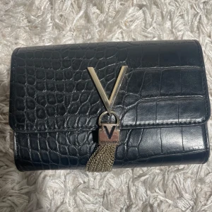 Svart kuvertväska från Valentino - Stilren svart kuvertväska från Valentino med krokodilpräglat skinn och snygga guldfärgade kedjedetaljer framtill. Väskan har ett stort V i metall och flera fack inuti, inklusive dragkedja. Perfekt för att lyfta din outfit med en lyxig touch.