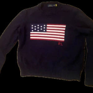 Mörkblå stickad tröja Polo Ralph Lauren - Mörkblå stickad tröja från Polo Ralph Lauren med amerikansk flagga broderad på bröstet och RL-detalj i rött. Den passar 160-176Klassisk rund halsringning och ribbade muddar. Perfekt för dig som vill sticka ut med en ikonisk look.