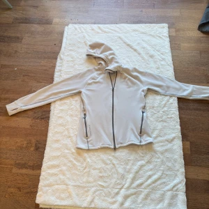 Beige fleecehoodie från Houdini - Säljer en beige hoodie från Houdini i mjuk fleece. Tröjan har hel dragkedja framtill, två fickor med dragkedja och en skön huva. Perfekt för kyliga dagar och outdoor-häng. Snygg och enkel design med diskret Houdini-logga på huvan och vid fickan.