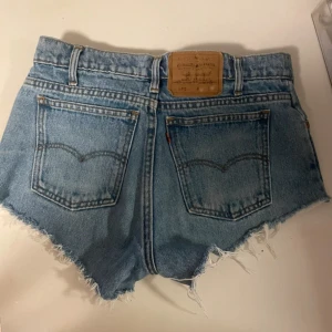 Levi's ljusblå jeansshorts med fransar - Säljer ett par klassiska ljusblå jeansshorts från Levi's med råa fransade kanter och femficksmodell. Shortsen har medel låg midja, bälteshällor och Levi's ikoniska läderpatch bak. Perfekta för sommaren och riktigt snygga till sneakers eller boots. Perfekt nu till halloween om man ska vara cowboy!🤠 