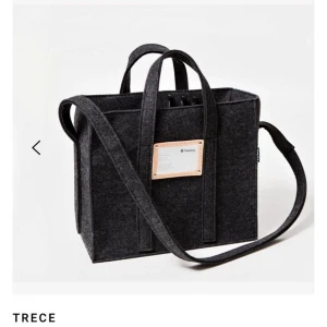 Svart datorväska i filt från Trece - Stilren svart datorväska från Trece i filtmaterial med både handtag och axelrem. Väskan har en fyrkantig form och en snygg detalj med en ljus etikett framtill. Perfekt för att bära laptop, pärmar och anteckningsblock.