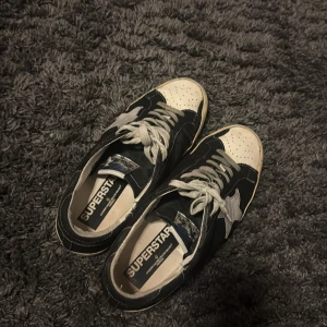 Golden Goose Superstar sneakers svart/vit - Golden Goose Superstar sneakers i svart och vitt med grå stjärndetalj på sidan. Skorna har snörning, perforerad tå och vit sula med vintage-look. Märkets logga syns tydligt på plösen och innersulan. Perfekta för dig som gillar streetstyle. 