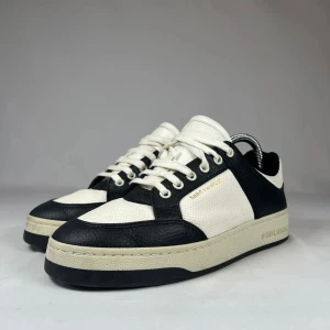 Saint Laurent svartvita sneakers i skinn - Snygga sneakers från Saint Laurent i svart och vitt skinn med perforerade paneler och klassisk snörning. Låg modell med rund tå och platt sula. Saint Laurent-logga i guld på sidan och präglad på sulan. Perfekt för dig som gillar stilrena och lyxiga sneakers.