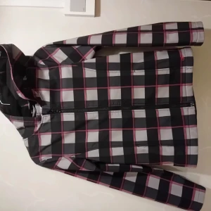 Rutig vindjacka med huva XL - Snygg vindjacka i svart, grått och vitt med rutor och röda linjer. Jackan har huva, hel dragkedja framtill och är helfodrad med mjuk fleece på insidan. Perfekt för blåsiga dagar och har en modern, sportig look.