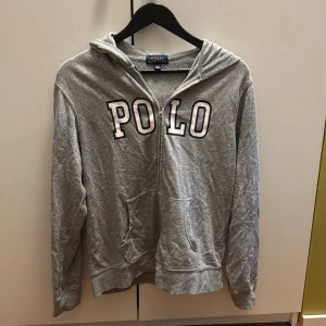 Grå hoodie från Polo Ralph Lauren - Grå hoodie från Polo Ralph Lauren med dragkedja och stor POLO-logga framtill. Tröjan har huva, känguruficka och långa ärmar. Perfekt för en chill och avslappnad stil.