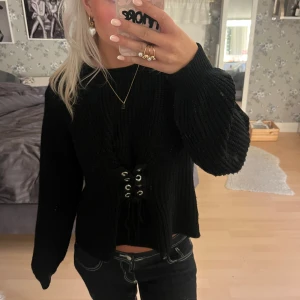 Svart stickad tröja med snörning - Svart stickad tröja med ballongärmar och snygg snörningsdetalj framtill i midjan. Tröjan har en lite loose passform och är perfekt för dig som gillar en trendig och bekväm look. Passar till både jeans och kjol för en cool vibe.