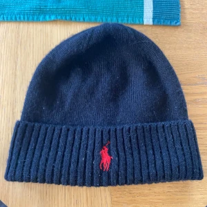 Svart merinoullsmössa från Polo Ralph Lauren - Svart ribbstickad mössa från Polo Ralph Lauren i 100% merinoull. Klassisk design med uppvikt kant och röd broderad logga framtill. Perfekt för kalla dagar och riktigt snygg till streetwear eller casual outfits.