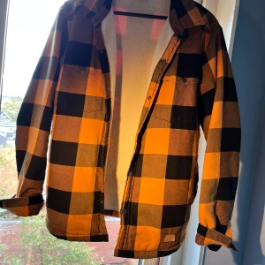 Rutig overshirt från Swedemount S - Overshirt från Swedemount i storlek S med stora orange och svarta rutor. Fodrad med mjuk fleece på insidan, två bröstfickor med knappar och klassisk krage. Perfekt lager-på-lager-plagg med avslappnad passform och snygg streetkänsla.