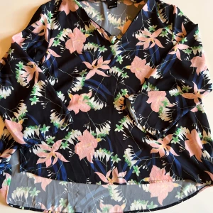 Blommig blus från Gina Tricot - Säljer en svart blus från Gina Tricot med vackert blommigt mönster i rosa, grönt och vitt. Blusen har v-ringning och långa ärmar, tillverkad i ett lätt och luftigt material som känns skönt mot huden. Perfekt för dig som gillar färg och mönster!