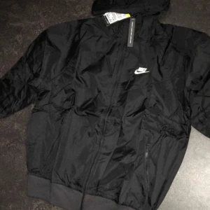Svart vindjacka från Nike - Svart vindjacka från Nike med klassisk logga på bröstet. Jackan har huva, dragkedja framtill och två sidofickor med dragkedja. Tillverkad i lätt och slitstark polyester, perfekt för blåsiga dagar. Enkel och stilren design som funkar till allt.