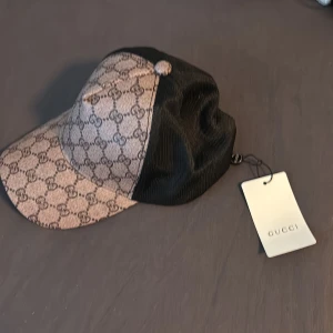 Gucci keps med GG-mönster och mesh - Snygg keps från Gucci med klassiskt GG-monogram i beige och brunt framtill och svart mesh baktill. Justerbar rem i svart skinn och broderad logga på insidan. Perfekt accessoar för att lyfta din streetwear-look.