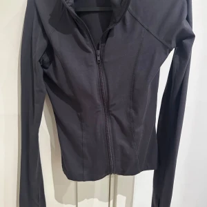 Grå zip hoodie med huva - Grå hoodie med dragkedja framtill och huva. Tröjan har lång ärm och en slim passform som sitter snyggt på kroppen. Perfekt för träning eller chill dagar. Materialet känns mjukt och stretchigt, troligen polyester eller bomullsmix.