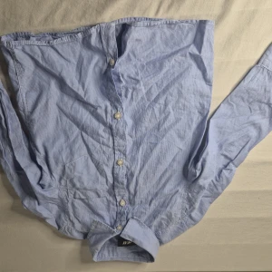 Blå randig skjorta från H&M barn - Snygg blå skjorta från H&M med tunna vita ränder och klassisk krage. Skjortan har långa ärmar, bröstficka och knäppning framtill. Perfekt för en stilren look till de små. Tillverkad i mjuk bomull för skön känsla hela dagen.