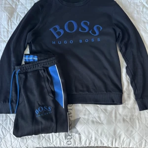 Hugo Boss tracksuit - Svart sweatshirt från Hugo Boss med blå logga och text på bröstet. Tröjan har långa ärmar och ribbade muddar vid ärmslut och nederkant. Materialet är mjukt och skönt, perfekt för en chill look. Matchande svarta byxor med blå och vit detalj på sidan ingår ej.