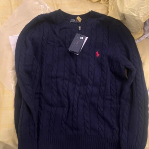 Mörkblå kabelstickad tröja Polo Ralph Lauren - Kabelstickad mörkblå eller svart  tröja från Polo Ralph Lauren med klassisk rund halsringning och röd broderad logga på bröstet. Tröjan är i mjuk bomull och har ribbade muddar vid ärmslut och nederkant. Perfekt för lager-på-lager under hösten och vintern.