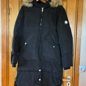 Svart lång pufferjacka med fuskpäls - Lång svart pufferjacka med huva och fuskpäls, dragkedja framtill och tryckknappar. Jackan har två stora fickor med lock och ribbade muddar vid ärmsluten. Perfekt för kalla vinterdagar och riktigt mysig att bära. En defekt se tredje bild!