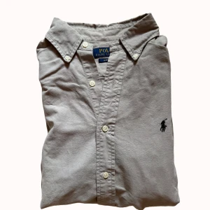 Grå långärmad skjorta Polo Ralph Lauren - Snygg grå skjorta från Polo Ralph Lauren med klassisk krage och broderad logga på bröstet. Långärmad modell med vita knappar framtill och vid ärmslut. Tillverkad i mjuk bomull och har en slim fit passform som sitter schysst.