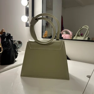 Grön handväska  - Snygg ljusgrön handväska från j med stilren, geometrisk form och guldfärgad logga. Väskan har ett unikt handtag som kan bäras runt handleden och är tillverkad i slätt skinn. Perfekt för dig som gillar trendiga accessoarer med minimalistisk vibe.