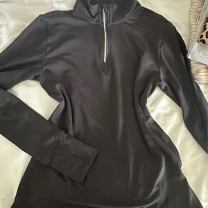 Svart långärmad träningströja half zip - Svart långärmad träningströja med half zip och hög krage. Tröjan har tumhål vid ärmsluten för extra komfort och är tillverkad i ett stretchigt syntetmaterial. Aldrig använd