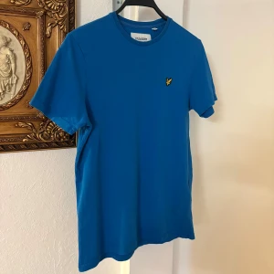 Blå t-shirt från Lyle & Scott - Snygg blå t-shirt från Lyle & Scott i ekologisk bomull. Klassisk passform med rund hals och korta ärmar. På bröstet sitter den ikoniska gula örn-loggan som brodyr. Perfekt för en clean och stilren look.