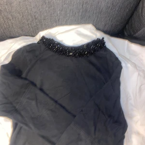 Svart långärmad tröja med pärlor - Svart långärmad tröja med snygga svarta pärlor runt halsen som ger en cool detalj. Tröjan har en dragkedja i ryggen och är gjord i ett mjukt material som känns skönt mot huden. Perfekt för dig som gillar att sticka ut med stilrena detaljer.