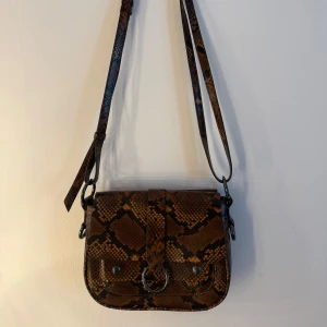 Axelväska Zadig & Voltaire i ormskinnsmönster - Snygg axelväska från Zadig & Voltaire med brunt och svart ormskinnsmönster. Väskan har en justerbar axelrem, silverfärgade detaljer och en metallring framtill. Perfekt storlek för det viktigaste och cool design med präglad logga på framsidan.